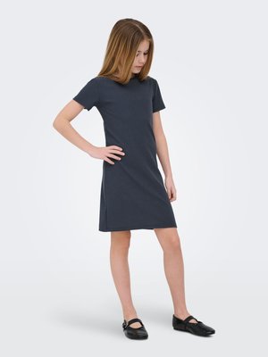 Pige med lyst, lige brunt hår iført en kortærmet mørk ribbet kjole og sorte Mary Jane-sko, stående med den ene hånd på hoften.