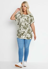 Top verde e crema con motivo a fiori e maniche a pipistrello, abbinato a jeans aderenti blu e sneakers bianche. Tessuto morbido con design floreale.