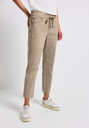 Beige casual broek met elastische tailleband en trekkoord, gedragen met witte sneakers door een staande persoon tegen een effen achtergrond.