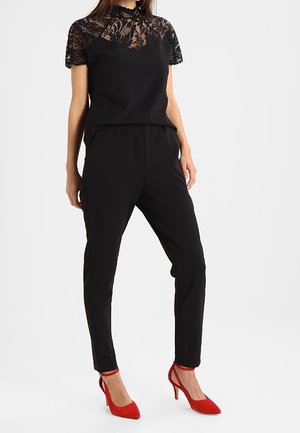 Femme portant un haut à manches courtes en dentelle noire, un pantalon noir et des chaussures à talons hauts rouges avec bride à la cheville, debout sur fond blanc.