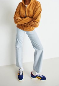 Veste zippée marron avec un col haut et un logo sur les manches, jeans droits bleu clair et baskets bleues avec des rayures jaunes et des lacets blancs.