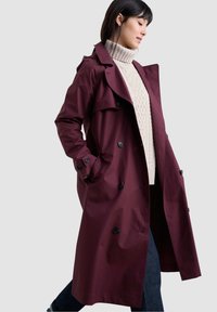 Bordeaux trenchcoat met een dubbelzijdige sluiting, zwarte knopen en zijzakken. Draag over een crèmekleurige, gebreide coltrui.