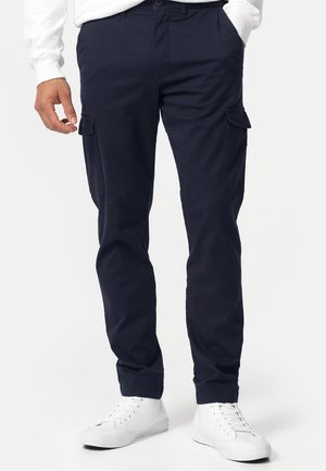 Homme portant un pantalon cargo bleu marine avec poches à rabat et des baskets blanches, debout avec une main dans la poche sur un fond uni.