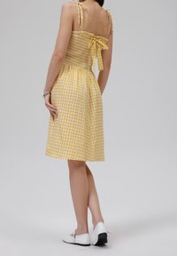 Žluté kostkované šaty s nařaseným korzátem, zavázacími ramínky a mašlí v pase. Sukně je po kolena s lehkým a splývavým materiálem.