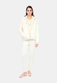 Giacca a vento color crema con design trapuntato, abbinata a pantaloni jogging crema e décolleté beige. Una collana in oro aggiunge un tocco discreto.