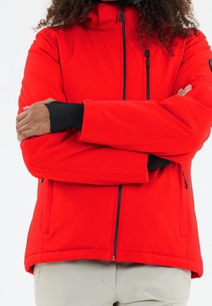Persoon met gekruiste armen, gekleed in een felrode jas met rits en lichtgrijze broek, zichtbaar van schouders tot heupen.