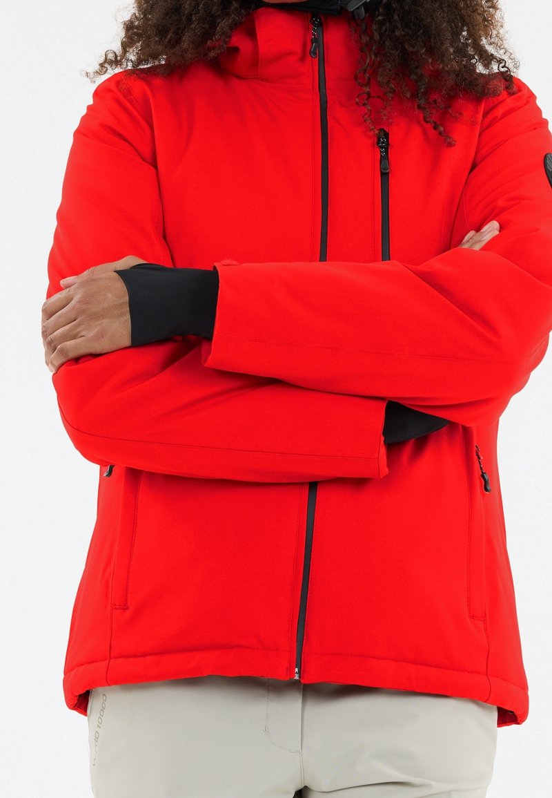 Personne les bras croisés portant une veste rouge vif zippée et un pantalon gris clair, montrée des épaules aux hanches.