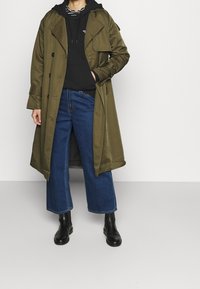 Olivgrön trenchcoat med dubbelknäppt design, kombinerad med en svart hoodie och vida blå jeans. Svarta ankelskor fulländar looken.