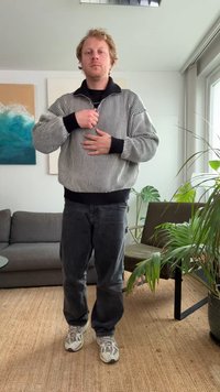 Fekete-fehér csíkos, túlméretezett pulóver magas gallérral, sötét farmerrel és világos tornacipővel párosítva, egy világos beltéri környezetben állva.