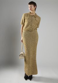 Guld sequined to-delt sæt med en kortærmet skjorte med krave og en tætsiddende nederdel. Model der holder en lille beige håndtaske og bærer sorte spidse sko.