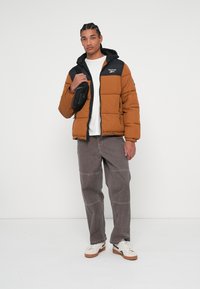 Casaco puffer em castanho com detalhes em preto, apresentando um fecho de correr e capuz. Combinado com calças cargo cinzas e ténis brancos.