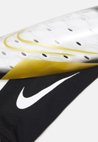 Nike skins her beskyttelse med et skinnende hvidt og sort design med hexagonale mønstre, guld accenter og en sort meshærme med et hvidt logo.