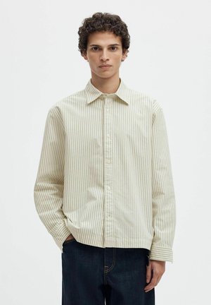 Jeune homme aux cheveux bouclés portant une chemise beige et blanche à rayures verticales et un jean bleu foncé sur un fond blanc uni.