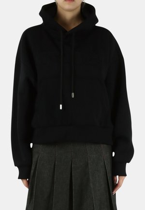 Persona che indossa una felpa nera con coulisse e una gonna plissettata scura, in piedi contro uno sfondo chiaro e uniforme.