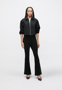 Μαύρο cropped μπουφάν με φερμουάρ μπροστά, σε συνδυασμό με εφαρμοστά μαύρα καμπανιστά παντελόνια και μωβ ψηλοτάκουνα, σε απλό λευκό φόντο.