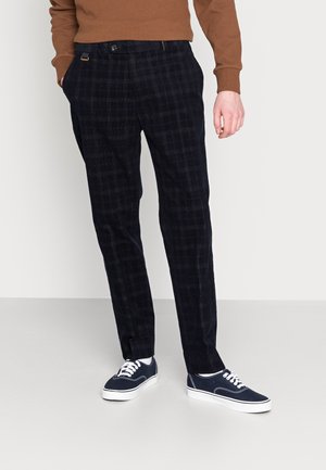 Marineblaue karierte Hose aus strukturiertem Stoff, mit Knopfverschluss und Taschen, kombiniert mit marineblauen Sneakers vor grauem Hintergrund.