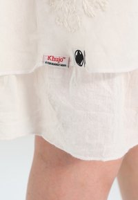 Weißer, transparenter Stoff mit zwei Kleidungsetiketten, einem rot-schwarzen „Khujo“-Markenetikett und einem schwarz-weißen Logotag, der in der Nähe eines Beins einer Person gezeigt wird.