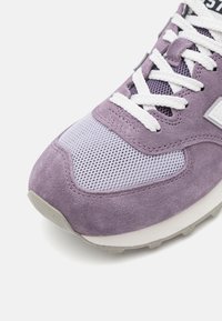 Fioletowy zamszowy sneaker z siateczkowymi akcentami, białymi sznurowadłami i gumową podeszwą. Charakterystyczne detale to fakturowane powierzchnie i subtelne różnice kolorystyczne.