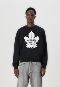 Hanorac negru cu un logo mare alb al echipei Toronto Maple Leafs, având un design cu frunze de arțar pe partea din față. Materialul pare moale, cu mansete cu nervuri.