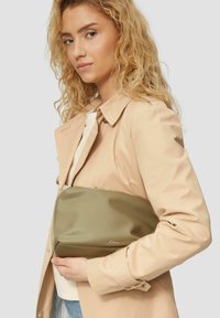 Zielona torebka kopertówka o gładkiej, błyszczącej fakturze, z zamkiem błyskawicznym i małym logo. Połączona z beżowym płaszczem i jasną koszulą.