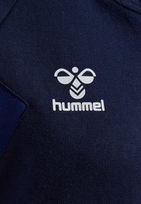 Białe stylizowane logo pszczoły i napis "hummel" na ciemnoniebieskiej teksturowanej tkaninie, prawdopodobnie odzież sportowa lub lekkoatletyczna.