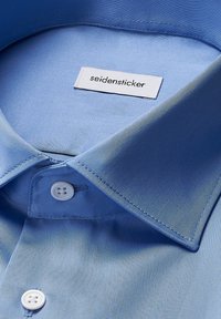 Lichtblauwe shirt met een gladde textuur, puntige kraag en witte knopen. Label met de tekst "seidensticker" is zichtbaar in de halsopening.