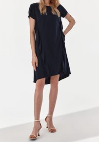 Navyblaues kurzärmeliges Kleid mit seitlichen Drapierungen, asymmetrischem Saum, aus leichtem Stoff, kombiniert mit beige hohen Sandalen.