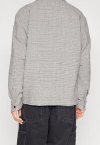 Giacca grigia con texture, colletto, polsini abbottonati e taglio dritto. Il materiale presenta un motivo intrecciato. Indossata sopra pantaloni scuri.