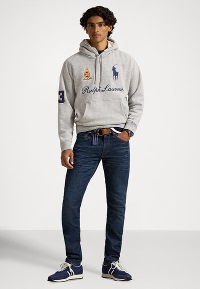 Młody mężczyzna stoi ubrany w jasnoszarą bluzę z kapturem Ralph Lauren, ciemnoniebieskie dżinsy, niebieskie trampki i zegarek, na tle jednolitego białego tła.