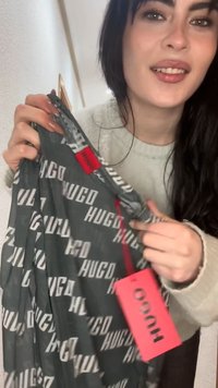 Γυναίκα που κρατάει ένα μαύρο ένδυμα με το μοτίβο Hugo και κόκκινη ταμπέλα, χαμογελώντας και δείχνοντας στην ετικέτα της μάρκας σε εσωτερικό χώρο.