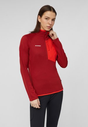 Giovane donna che indossa una giacca rossa Mammut con zip, tasca arancione sul petto e pantaloni neri su sfondo grigio uniforme.