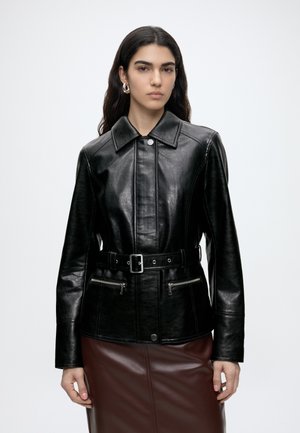Donna che indossa una giacca di pelle nera con cintura e tasche con zip e una gonna di pelle marrone scuro, in piedi davanti a uno sfondo bianco uniforme.