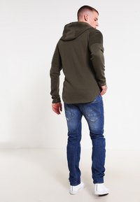 Felpa con cappuccio verde oliva con dettagli a coste sui gomiti, abbinata a jeans in denim blu e sneakers bianche.