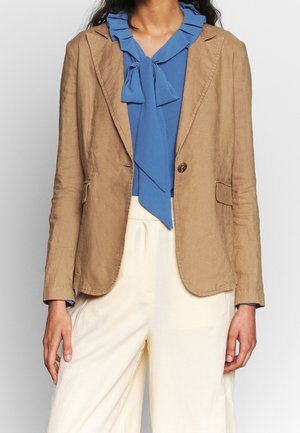 Lysebrun linned blazer med klippede revers, enkelt knaplukning og to forlommer, parret med en blå rynket bluse og hvide bukser.