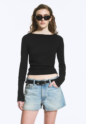 Kobieta o falowanych włosach, ubrana w czarny krótki top z długim rękawem, jasne krótkie spodenki jeansowe z czarnym paskiem i czarne okulary przeciwsłoneczne w stylu kocich oczu, stoi z ręką w kieszeni.