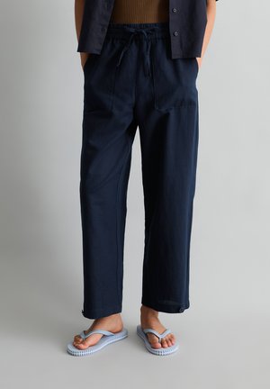 Persoon dragen marineblauwe broek met trekkoord en voorzakken en lichtblauwe slippers, staand tegen een effen grijze achtergrond.