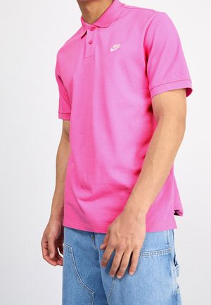 Mann trägt ein knallpinkes Nike-Poloshirt und hellblaue Jeans, steht vor einem schlichten weißen Hintergrund.