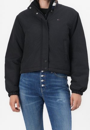Modèle portant une veste noire à boutons-pression, avec un col relevé et des poignets élastiqués, associée à un jean bleu à braguette boutonnée.