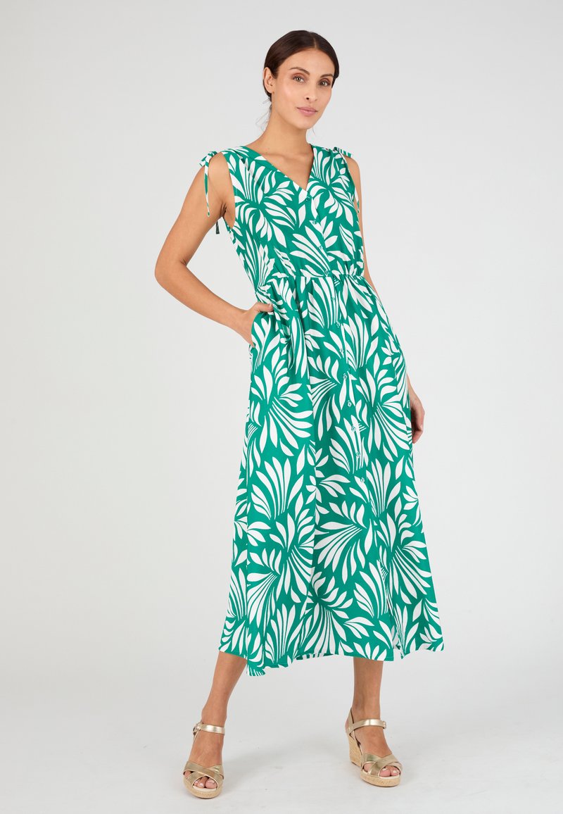 Femme portant une robe midi verte sans manches avec des motifs de feuilles blanches et des bretelles à nouer aux épaules, posant les mains dans les poches et des sandales compensées dorées.