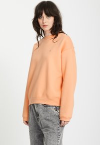 Sweat-shirt de couleur pêche avec col rond, épaules tombantes et ourlet côtelé. Associé à un pantalon gris texturé. Détail de logo simple sur la poitrine.