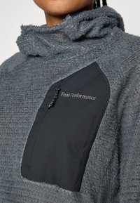 Pull-over en polaire gris avec un extérieur texturé, doté d'une poche poitrine zippée noire et du logo "Peak Performance" brodé en argent.
