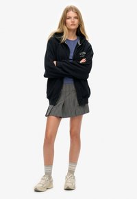 Sort zip-up hoodie med hvide kraveaccentuer, lagdelt over en lilla skjorte. Grå stribet plisseret nederdel, grå og beige sneakers, og stribede sokker.