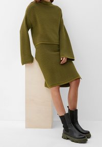 Ensemble pull et jupe en tricot rayé vert olive avec manches longues et texture côtelée, associé à des bottines noires avec semelles en caoutchouc vertes.