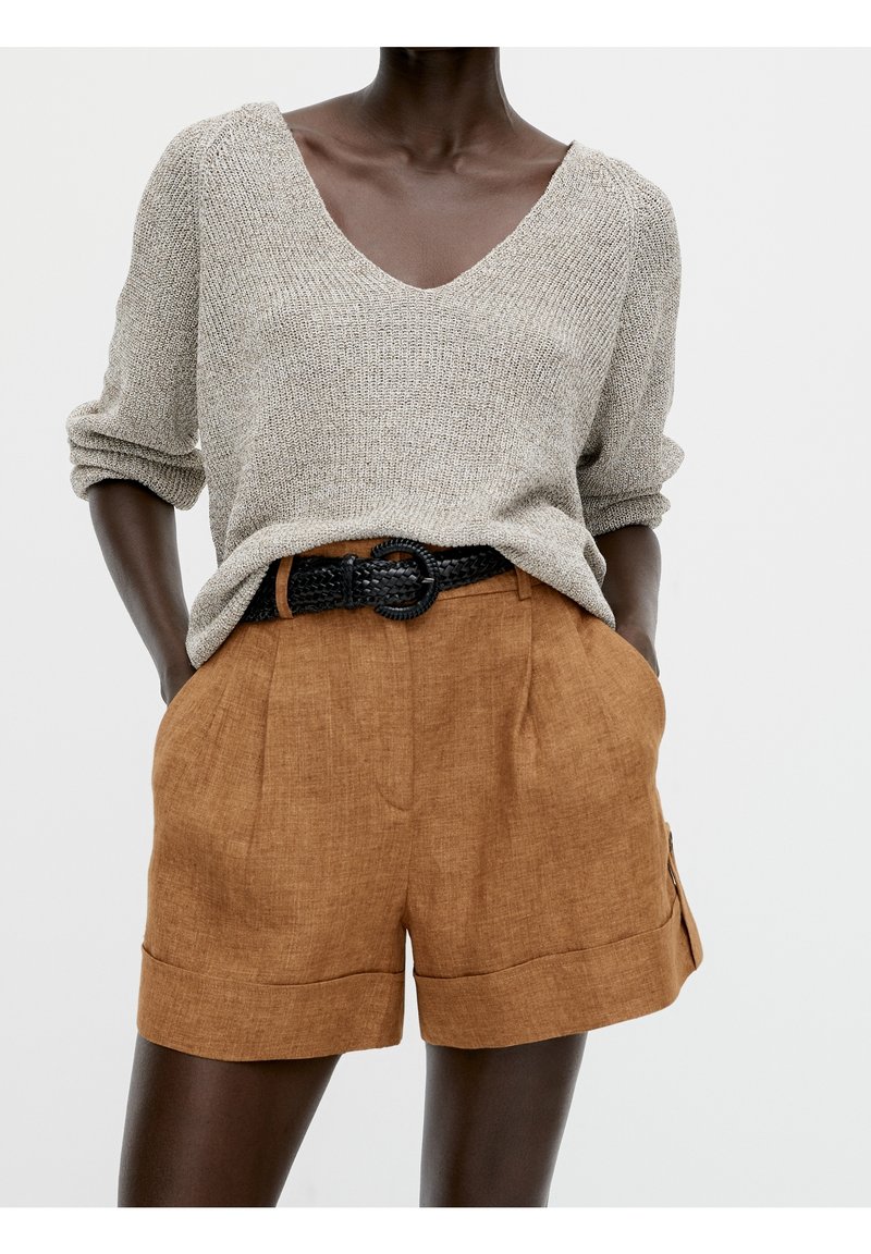 Pull en maille gris clair avec un col en V, associé à un short en lin marron avec un ourlet roulé et une ceinture tressée noire.