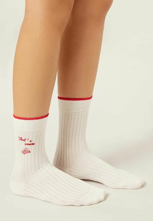 Gambe con calzini alla caviglia bianchi a coste con bordo rosso e testo ricamato rosso e motivo sul lato esterno del calzino sinistro.