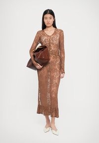 Vestido de renda em castanho, com mangas longas, decote em V e uma bainha com babados. Modelo segura uma mala de couro castanha com um detalhe dourado.