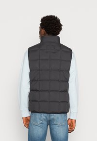 Gilet imbottito nero con colletto alto e design trapuntato. Indossato sopra una camicia a maniche lunghe blu chiaro, con una texture liscia e opaca.