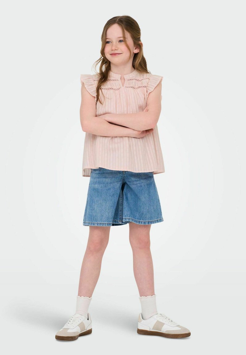 Ljusrosa, ärmlös topp med volangaxlar och vertikala ränder; matchad med blekta denimshorts; vita sneakers med beige detaljer.