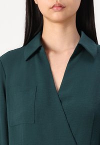 Zöldeskék, textúrázott blúz, hegyes gallérral és bal oldalon egy mellzsebbel.