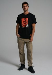 Czarny t-shirt z czerwoną grafiką przedstawiającą napis "Dakar", połączony z beżowymi spodniami cargo. Czarne obuwie dopełnia strój.